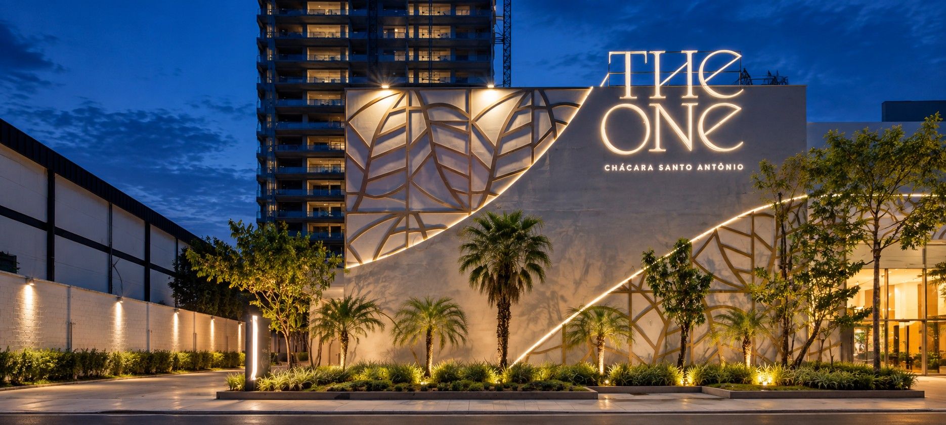 Estande The One, da Cury, ao anoitecer, com painel vazado de folhas, letreiro The One iluminado e linha de LED na fachada