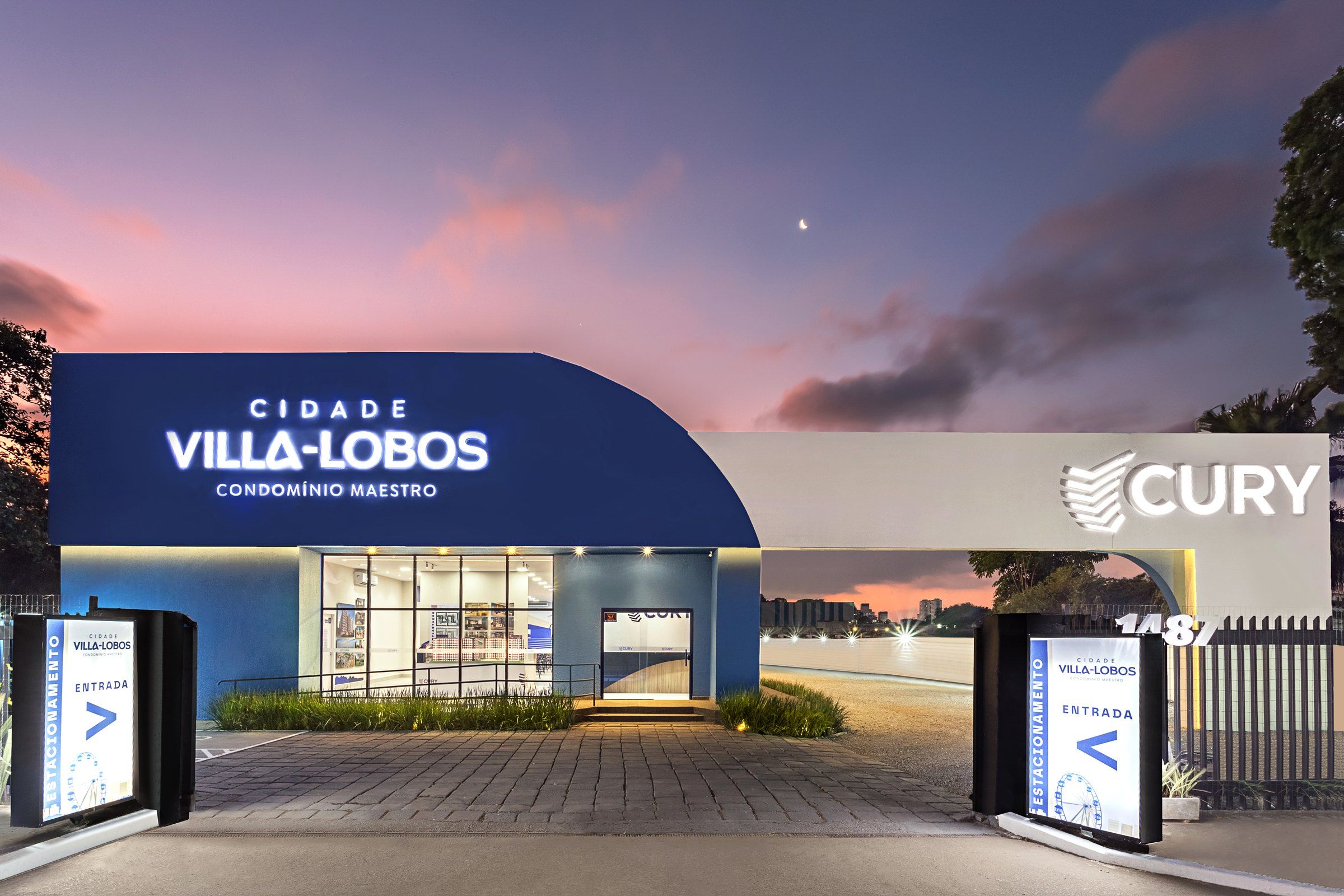 Estande de vendas Cidade Villa-Lobos, da Cury, ao entardecer, com letreiro iluminado e totens luminosos de entrada